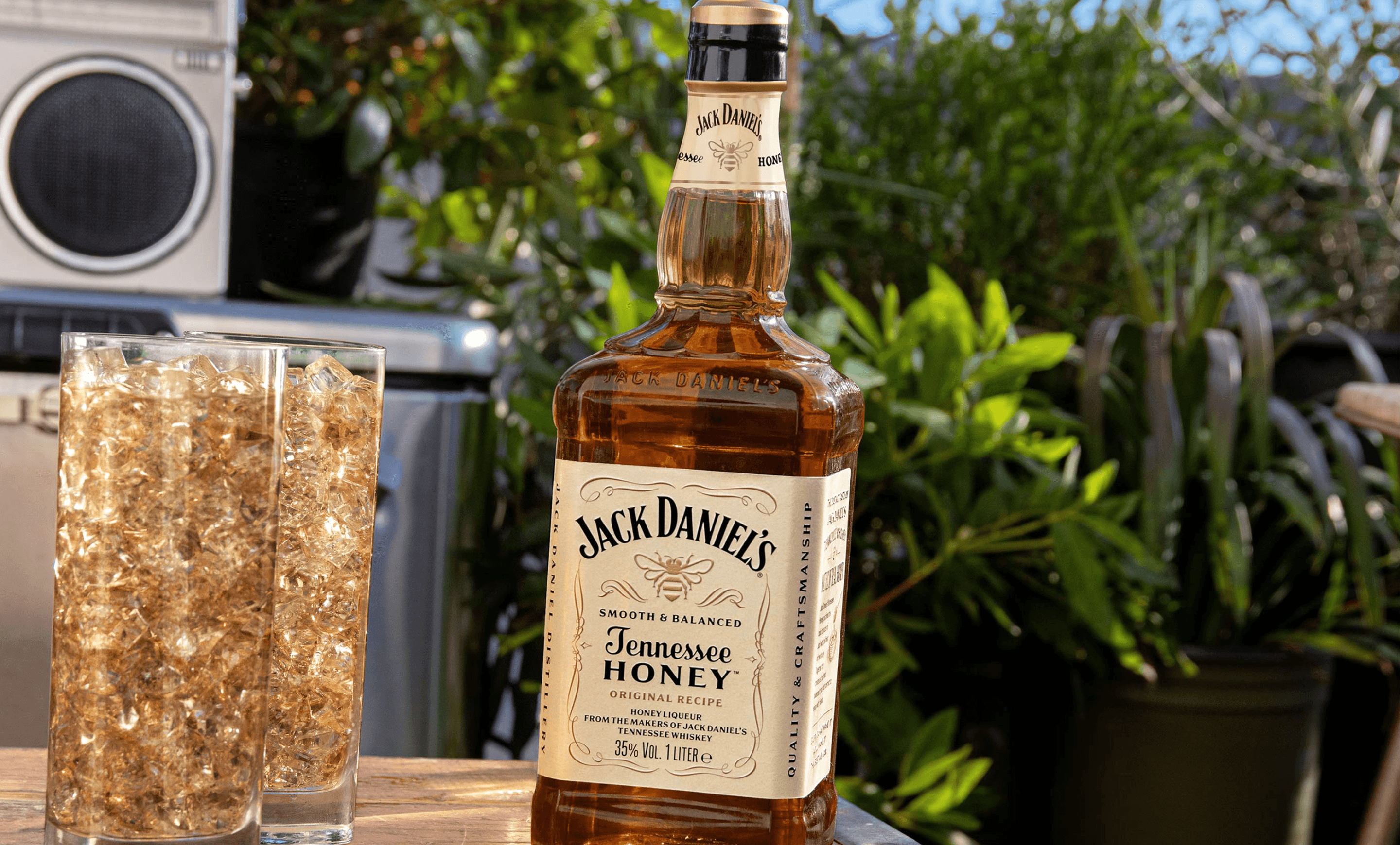 Picture of the Jack Daniel's Tennessee Honey Bottle in a corn field next to 2 tall glasses fill with ice and a cocktail